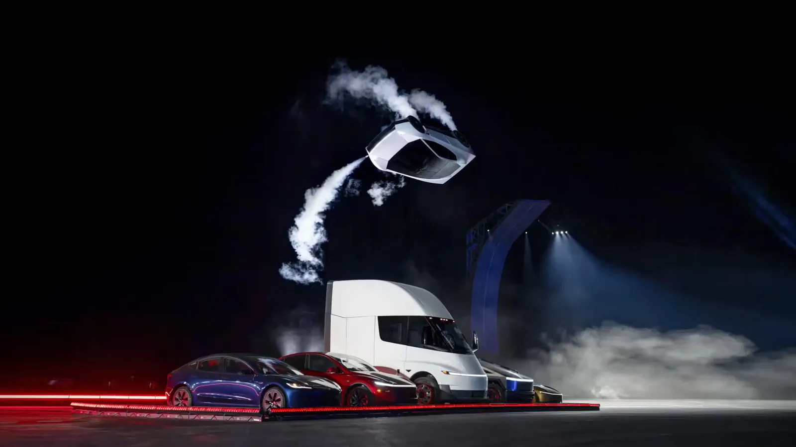 A white car flipping upside down in mid-air, with trails of white gas streaming behind it, above a row of parked Tesla vehicles including a Semi truck, on a dark stage with blue and red lighting.
